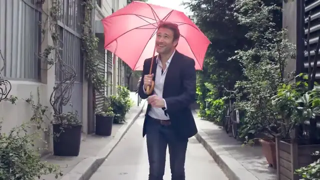 Un homme en costume sourit sous un parapluie rose dans une ruelle parisienne